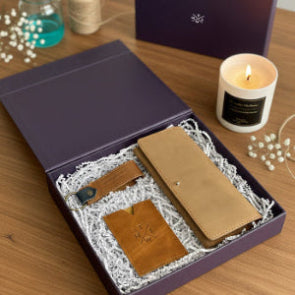 Brown leather wallet and cardholder set in a gift box with a candle keychain and decorative items on a wooden surface.