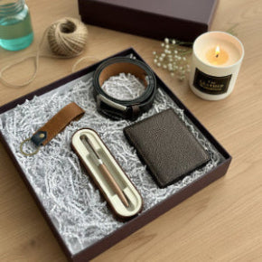 Gift set with leather belt, wallet, pen, and keychain in a box on a wooden surface with a candle.