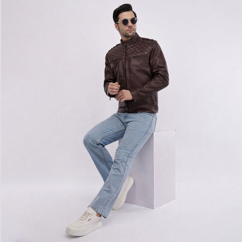 Man wearing a brown leather jacket and light blue jeans sitting on a white cube against a white background