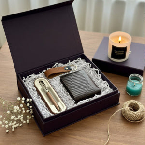 Gift set with wallet, pen, and keychain in an open box on a wooden surface with candles and yarn.