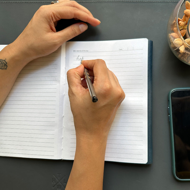Person writing in a notebook with a pen on a dark surface, next to a phone and small container.