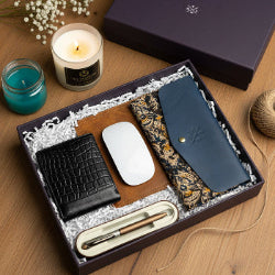 Gift set with black wallet, white mouse, blue phone case, and pen in a box on a wooden surface.