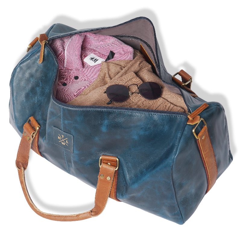 Blue duffel bag with brown leather straps containing clothes and sunglasses on a white background
