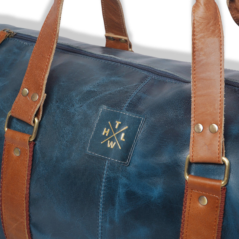Blue leather duffel bag with brown straps and a logo on a white background