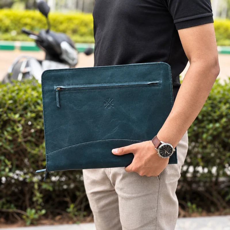 Man in a black polo and beige trousers holding a teal blue leather portfolio with a front zip pocket and embossed logo.