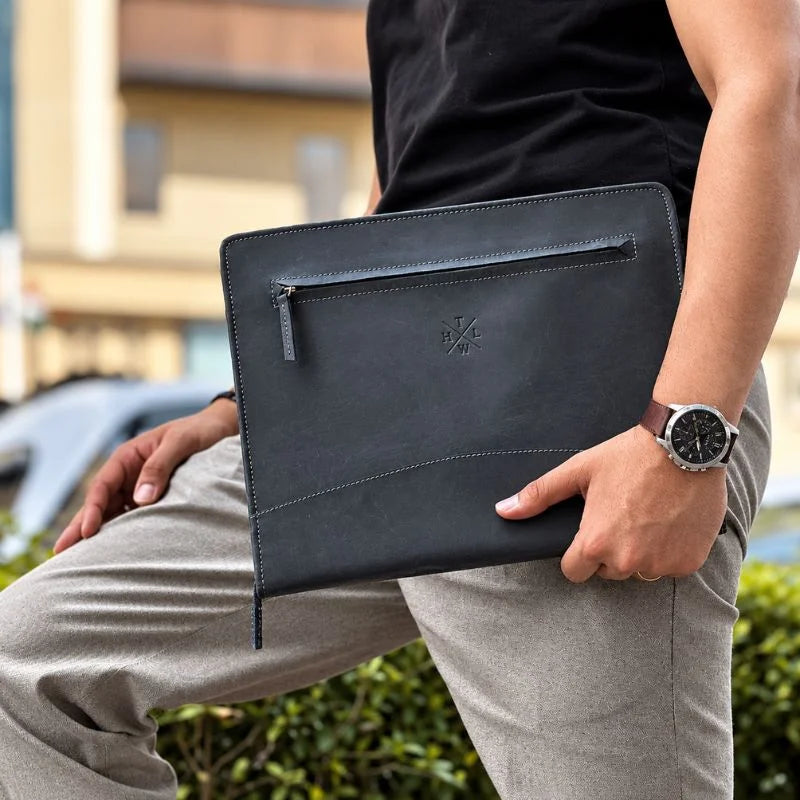 Man holding a charcoal grey leather portfolio with a front zip pocket and embossed logo.