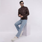 Man wearing a brown leather jacket and light blue jeans sitting on a white cube against a white background