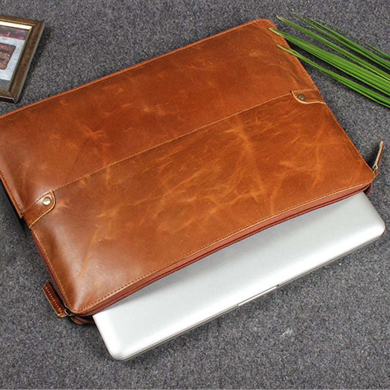 Premium brown leather laptop sleeve with a zipper, shown partially open with a silver laptop inside, placed on a grey fabric surface.