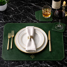 Dining table setting with green placemat and coaster white plates, gold cutlery, and glasses on a dark marble surface.