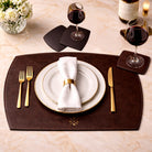 Dining table setting with white plates, gold cutlery, and a brown placemat and coaster on a marble surface.