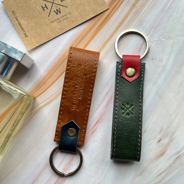 Two leather keychains on a light wooden surface with a perfume bottle and card in the background.