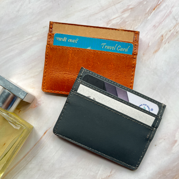 Two leather card holders, one brown and one black, on a light marble surface with a travel card and perfume bottle.