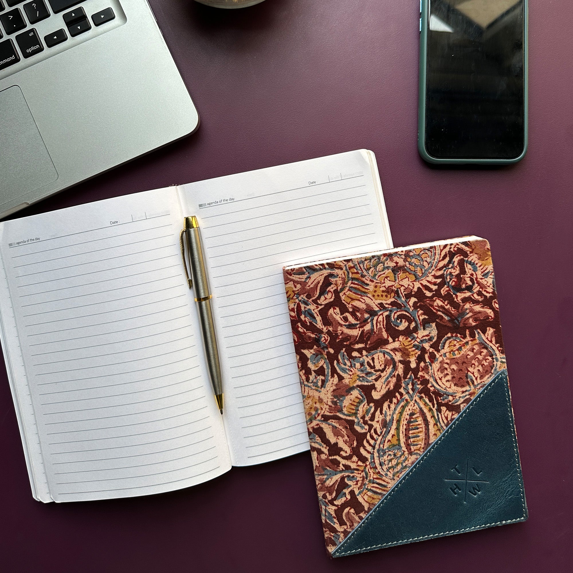 Open notebook with a pen, decorative notebook, laptop, and phone on a dark surface