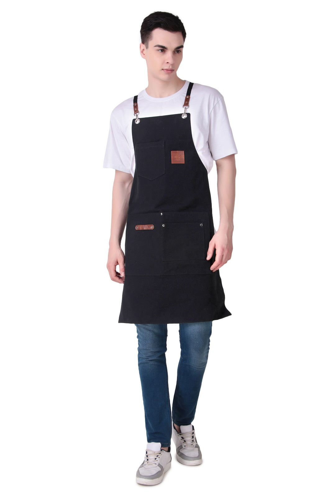 A man modeling a black bib apron with brown leather-like accents, worn over a white t-shirt and blue jeans, against a plain white background.