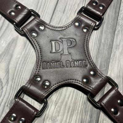 Brown leather harness with studded details on a wooden surface, featuring the brand 'DP' by Daniel Pange.