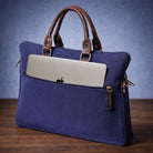 Blue canvas laptop bag with brown handles holding a silver laptop on a wooden surface with a blue background