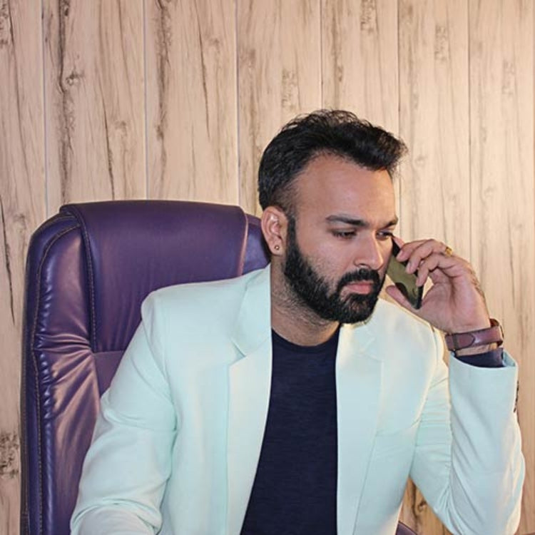 Man in a light blue blazer sitting in a purple chair, talking on a phone with a wooden wall background.