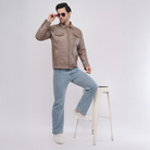 Man wearing a brown leather jacket and light blue jeans, standing on a white stool against a plain background.