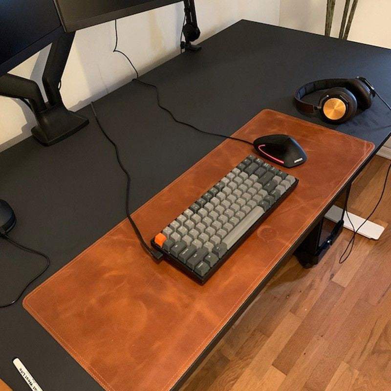 A black desk setup featuring a brown leather desk mat with a compact mechanical keyboard, a wired mouse, and a pair of over-ear headphones placed on the right side.