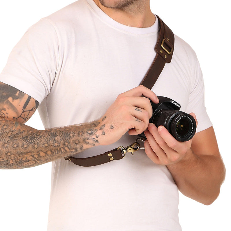 A man wearing a white t-shirt and a brown leather camera strap is holding a Canon DSLR camera; the man has a full sleeve tattoo on his left arm.