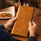 Person holding a brown leather menu with 'ARAK' embossed, on a wooden table with food and drinks.