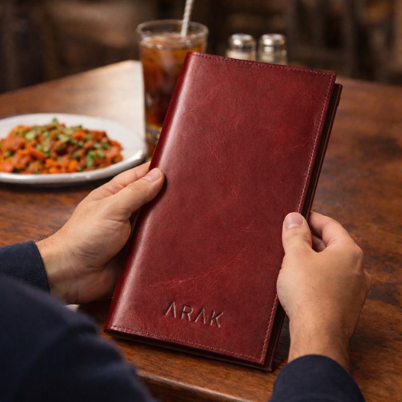 Person holding a red leather menu with 'ARAK' branding on a table with food and drinks.