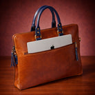 Brown leather handbag with blue handles holding an Apple laptop on a wooden surface.