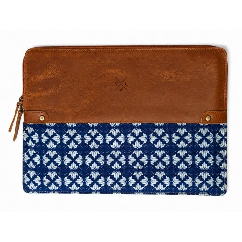 A zip-around pouch featuring a smooth brown leather top half and a navy blue fabric bottom half with a white geometric ikat pattern. The pouch has brass grommets, a leather zipper pull, and an embossed logo on the leather section.