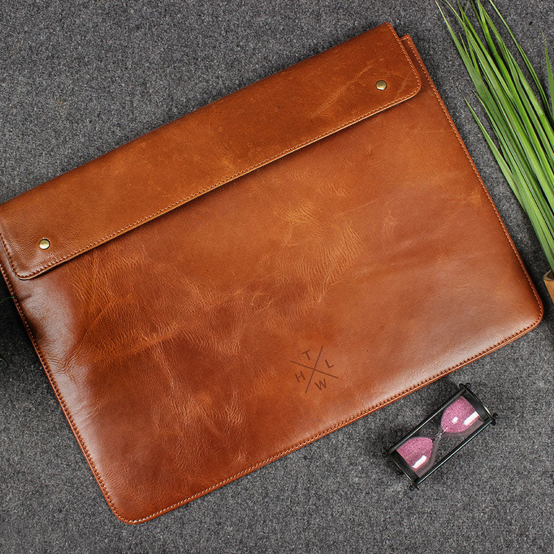 Premium brown leather laptop sleeve with flap closure placed on a grey surface beside a small hourglass and green leaves