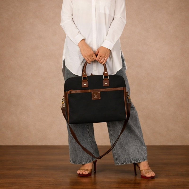 Person holding a black canvas handbag with brown straps against a beige background