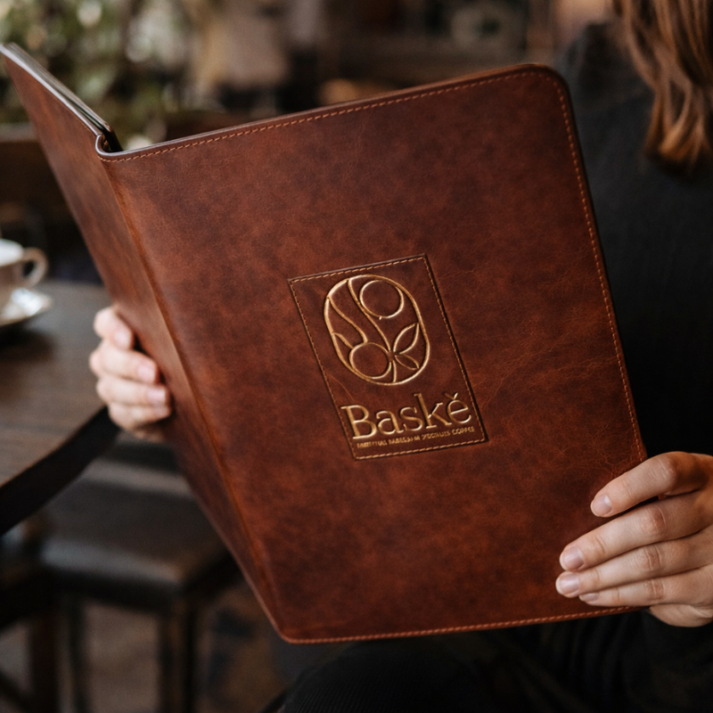 Person holding a brown menu with 'Baské' logo in a blurred cafe setting