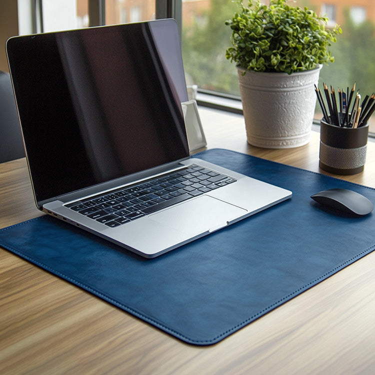 A sleek laptop placed on a blue leather desk mat beside a wireless mouse on a wooden desk, with a potted plant and pencil holder near a window in the background.