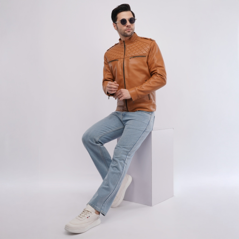 Man in brown leather jacket and light blue jeans sitting on white cube on white background.